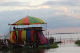 A colorful, multi-colored tent structure sits beside a large body of water, likely a river or lake. The tent is elevated on a platform and supported by scaffolding. People are gathered underneath and around the tent structure, with some sitting on the grass nearby. The sky is partially cloudy, with soft light suggesting it is either early morning or late afternoon.