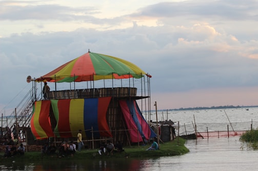 A colorful, multi-colored tent structure sits beside a large body of water, likely a river or lake. The tent is elevated on a platform and supported by scaffolding. People are gathered underneath and around the tent structure, with some sitting on the grass nearby. The sky is partially cloudy, with soft light suggesting it is either early morning or late afternoon.