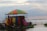 A colorful, multi-colored tent structure sits beside a large body of water, likely a river or lake. The tent is elevated on a platform and supported by scaffolding. People are gathered underneath and around the tent structure, with some sitting on the grass nearby. The sky is partially cloudy, with soft light suggesting it is either early morning or late afternoon.
