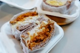 A close-up of two packaged baked goods placed on white disposable trays. Each item appears to be a type of cake or bread filled with a creamy substance and topped with a layer of golden-brown crumbs. The surface texture looks soft and gooey, highlighting the richness of the filling. The items are partially wrapped in printed paper, giving a rustic and homemade feel.