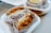 A close-up of two packaged baked goods placed on white disposable trays. Each item appears to be a type of cake or bread filled with a creamy substance and topped with a layer of golden-brown crumbs. The surface texture looks soft and gooey, highlighting the richness of the filling. The items are partially wrapped in printed paper, giving a rustic and homemade feel.