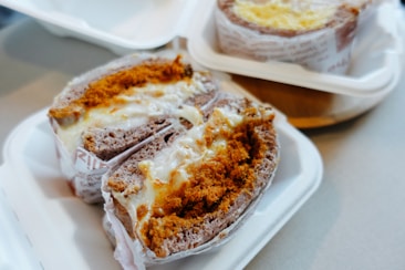 A close-up of two packaged baked goods placed on white disposable trays. Each item appears to be a type of cake or bread filled with a creamy substance and topped with a layer of golden-brown crumbs. The surface texture looks soft and gooey, highlighting the richness of the filling. The items are partially wrapped in printed paper, giving a rustic and homemade feel.