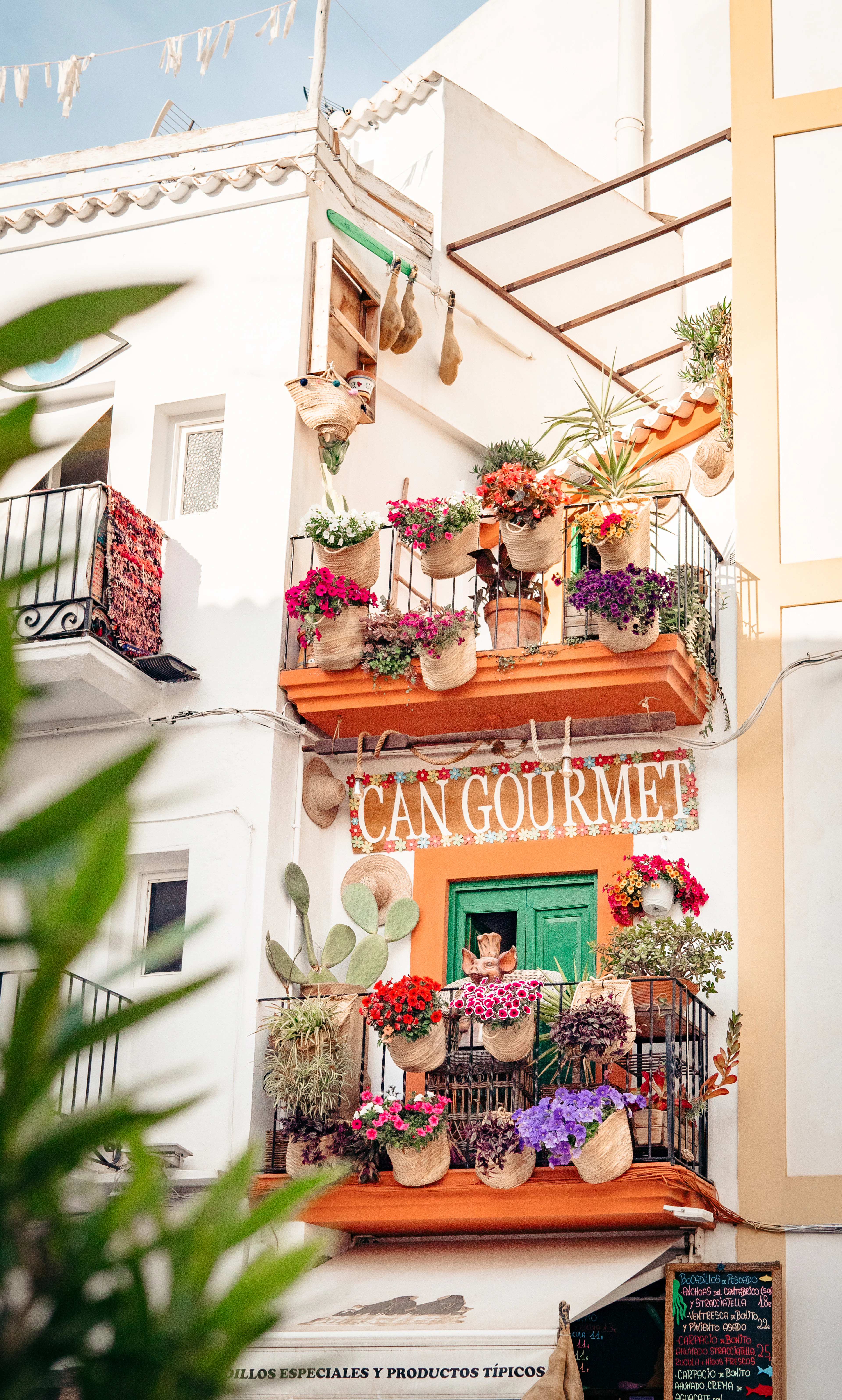 A flower-covered balcony spills geraniums over the 'Can Gourmet' shopfront on a whitewashed Ibiza Town street.
