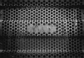An intricate pattern of hexagonal holes forms a metal grate, likely used for fencing or as an industrial barrier. The surface appears worn and rusted, conveying a sense of age and industrial use.