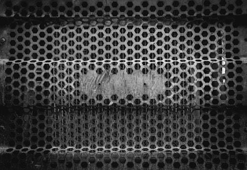 An intricate pattern of hexagonal holes forms a metal grate, likely used for fencing or as an industrial barrier. The surface appears worn and rusted, conveying a sense of age and industrial use.