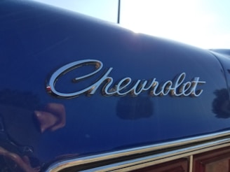 An angled view of a vintage car with the Chevrolet logo in chrome lettering on a shiny, dark blue surface. Reflections can be seen in the polished surface of the car.