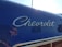 An angled view of a vintage car with the Chevrolet logo in chrome lettering on a shiny, dark blue surface. Reflections can be seen in the polished surface of the car.