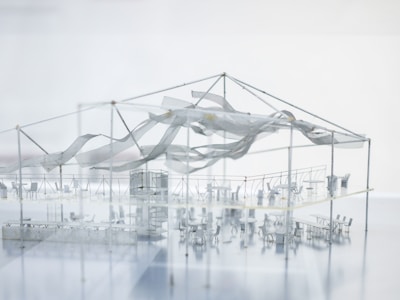A highly detailed architectural model showcases an indoor setup with various tables and chairs arranged in sections. The structure has a metal framework supporting a semi-transparent fabric that appears to be waving or draped artistically. There is a prominent central structure, possibly representing a stage or performance area. The overall impression is sleek and modern, with a minimalist design.