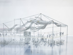 A highly detailed architectural model showcases an indoor setup with various tables and chairs arranged in sections. The structure has a metal framework supporting a semi-transparent fabric that appears to be waving or draped artistically. There is a prominent central structure, possibly representing a stage or performance area. The overall impression is sleek and modern, with a minimalist design.