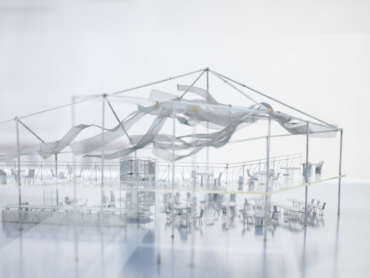 A highly detailed architectural model showcases an indoor setup with various tables and chairs arranged in sections. The structure has a metal framework supporting a semi-transparent fabric that appears to be waving or draped artistically. There is a prominent central structure, possibly representing a stage or performance area. The overall impression is sleek and modern, with a minimalist design.