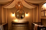 A cozy nursery features an elegant design, highlighted by a large round mirror above a wooden dresser, flanked by two wall lamps with a warm glow. A unique spherical chandelier hangs from the ceiling, contributing to the room's soothing ambiance. The space is enclosed with flowing, draped fabric against the walls, and framed animal prints hang on the right wall. A crib is situated on either side of the dresser, adding to the nursery setting.