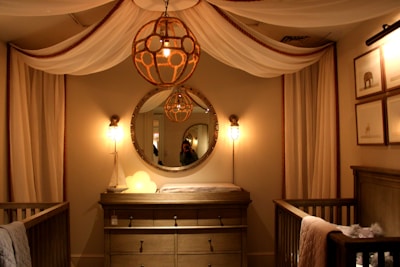 A cozy nursery features an elegant design, highlighted by a large round mirror above a wooden dresser, flanked by two wall lamps with a warm glow. A unique spherical chandelier hangs from the ceiling, contributing to the room's soothing ambiance. The space is enclosed with flowing, draped fabric against the walls, and framed animal prints hang on the right wall. A crib is situated on either side of the dresser, adding to the nursery setting.