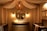 A cozy nursery features an elegant design, highlighted by a large round mirror above a wooden dresser, flanked by two wall lamps with a warm glow. A unique spherical chandelier hangs from the ceiling, contributing to the room's soothing ambiance. The space is enclosed with flowing, draped fabric against the walls, and framed animal prints hang on the right wall. A crib is situated on either side of the dresser, adding to the nursery setting.