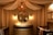A cozy nursery features an elegant design, highlighted by a large round mirror above a wooden dresser, flanked by two wall lamps with a warm glow. A unique spherical chandelier hangs from the ceiling, contributing to the room's soothing ambiance. The space is enclosed with flowing, draped fabric against the walls, and framed animal prints hang on the right wall. A crib is situated on either side of the dresser, adding to the nursery setting.