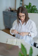 A person with glasses is seated at a desk with an open planner. They wear a white shirt and hold a pen in both hands. The setting includes a laptop, some indoor plants, and blurred office furniture in the background, suggesting a workspace environment.
