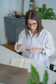 A person with glasses is seated at a desk with an open planner. They wear a white shirt and hold a pen in both hands. The setting includes a laptop, some indoor plants, and blurred office furniture in the background, suggesting a workspace environment.
