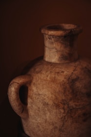 A weathered clay urn with a single handle, showcasing a textured surface with visible cracks and a rich earthy color palette. The lighting highlights the urn's aged appearance and intricate surface details.