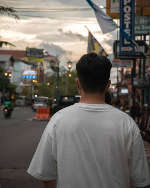 A person wearing a white t-shirt is seen from behind, walking down a city street lined with various signboards and shops. The sky appears cloudy as the sun sets, casting a warm, dim light on the surroundings. There are vehicles and street lights visible in the distance.