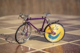 A miniature bicycle with a purple frame and intricate metallic detailing is placed on a tiled floor. The front wheel is traditional with spokes, while the back wheel features a decorative disk with a yellow and blue design, alongside some text.