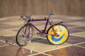 A miniature bicycle with a purple frame and intricate metallic detailing is placed on a tiled floor. The front wheel is traditional with spokes, while the back wheel features a decorative disk with a yellow and blue design, alongside some text.