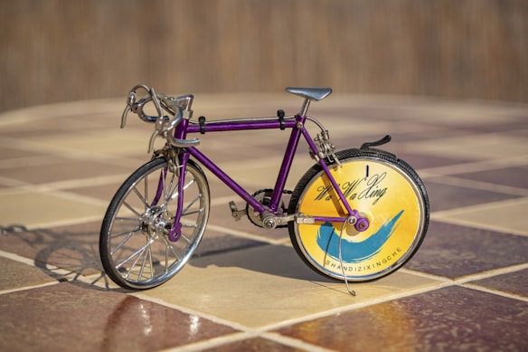 A miniature bicycle with a purple frame and intricate metallic detailing is placed on a tiled floor. The front wheel is traditional with spokes, while the back wheel features a decorative disk with a yellow and blue design, alongside some text.