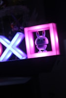 A cute figurine of a bunny is illuminated in a brightly colored frame. To the left of the bunny, there are neon shapes of an X and O, glowing in blue and pink hues. The background is dark, making the illuminated objects stand out vividly.