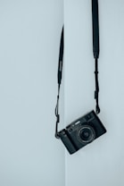 A black camera with a strap is hanging against a light blue wall. The camera is positioned at an angle, and the scene conveys a minimalist aesthetic.