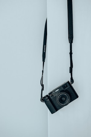 A black camera with a strap is hanging against a light blue wall. The camera is positioned at an angle, and the scene conveys a minimalist aesthetic.