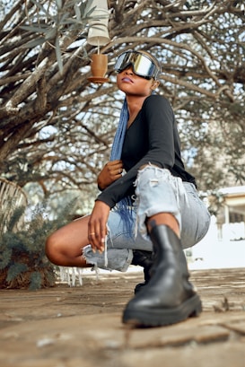 A person crouching outdoors, wearing futuristic sunglasses and a black long-sleeve shirt. They have long blue braids and are dressed in ripped denim shorts and black boots. The scene features a natural setting with intertwined branches in the background, creating an artistic and urban vibe.