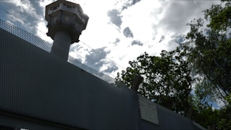 A tall watchtower rises above a high, textured metal fence, outlined against a cloudy sky. Barbed wire runs along the top of the fence, suggesting security or restricted access. Trees with dense foliage are visible nearby, contrasting with the man-made structures.