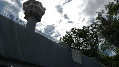 A tall watchtower rises above a high, textured metal fence, outlined against a cloudy sky. Barbed wire runs along the top of the fence, suggesting security or restricted access. Trees with dense foliage are visible nearby, contrasting with the man-made structures.