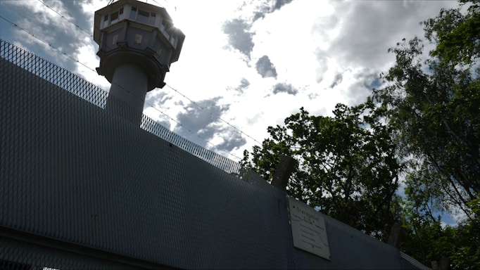A tall watchtower rises above a high, textured metal fence, outlined against a cloudy sky. Barbed wire runs along the top of the fence, suggesting security or restricted access. Trees with dense foliage are visible nearby, contrasting with the man-made structures.