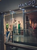 A display of mannequins dressed in elegant clothing is visible inside a brightly lit retail store. The store has a modern design with glass windows and a sleek metal railing. The clothing includes a green dress, a patterned outfit, and a white suit. The store's name, MANGO, is prominently displayed above the entrance.