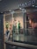 A display of mannequins dressed in elegant clothing is visible inside a brightly lit retail store. The store has a modern design with glass windows and a sleek metal railing. The clothing includes a green dress, a patterned outfit, and a white suit. The store's name, MANGO, is prominently displayed above the entrance.