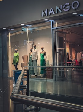 A display of mannequins dressed in elegant clothing is visible inside a brightly lit retail store. The store has a modern design with glass windows and a sleek metal railing. The clothing includes a green dress, a patterned outfit, and a white suit. The store's name, MANGO, is prominently displayed above the entrance.