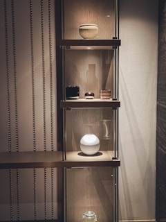 A stylish interior setup featuring a wooden shelf with multiple levels, displaying decorative items. The top shelf holds a round, ribbed vase. The middle shelf contains a black square object, a small round dish, and a striped container. Below, a spherical white vase is illuminated, creating a focal point. The backdrop has vertical metallic strips adding texture and visual interest.
