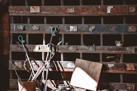 An old wooden shelf structure filled with various metal boxes and compartments. The shelves are weathered and aged, with labels and handles on some of the compartments. In front of the shelf, several metal objects including rods and a bucket are visible, casting shadows across the scene. Sunlight highlights a board leaning against the shelf, adding contrast and depth.