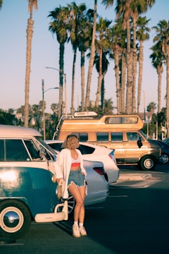 A person with curly hair stands near a vintage blue and white van, with a background of tall palm trees and several other parked vehicles, including a tan RV. The person is wearing a white shirt over a red and white striped top, denim shorts, and white footwear, and is holding a small bag.