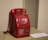 A vintage rotary dial payphone with a red color, featuring a coin slot and instructions displayed on a metal plate. It sits on a light-colored surface, with a coiled cord connecting the handset to the main body.