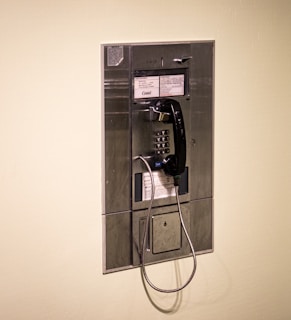 A wall-mounted payphone with a black handset attached by a coiled metal cord. The payphone has a metal surface with buttons for dialing numbers and instructions written above and below the keypad.