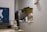 A tidy kitchen corner with a silver refrigerator, white cabinets, and various kitchen appliances on the countertop, including a kettle and a dish rack. There is a blue wall hanging featuring a white bear and stars on one side, and a small festive decoration hanging on the cabinet door.
