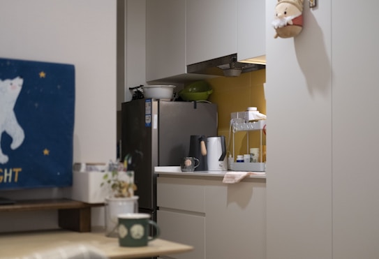 A tidy kitchen corner with a silver refrigerator, white cabinets, and various kitchen appliances on the countertop, including a kettle and a dish rack. There is a blue wall hanging featuring a white bear and stars on one side, and a small festive decoration hanging on the cabinet door.