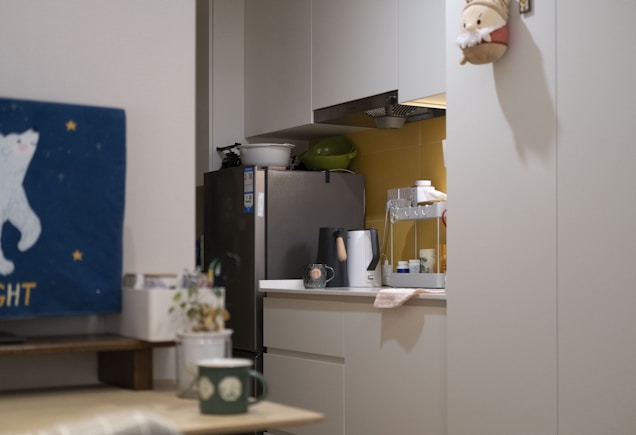 A tidy kitchen corner with a silver refrigerator, white cabinets, and various kitchen appliances on the countertop, including a kettle and a dish rack. There is a blue wall hanging featuring a white bear and stars on one side, and a small festive decoration hanging on the cabinet door.