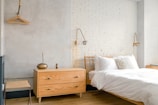 A bright and minimalist bedroom with light-colored wooden furniture, including a bed and a chest of drawers. The walls are painted in soft gray and white brick, creating a calming atmosphere. Two modern wall-mounted lamps provide light, and a simple wooden coat hanger is visible on one wall.
