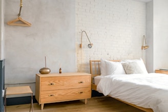 A bright and minimalist bedroom with light-colored wooden furniture, including a bed and a chest of drawers. The walls are painted in soft gray and white brick, creating a calming atmosphere. Two modern wall-mounted lamps provide light, and a simple wooden coat hanger is visible on one wall.