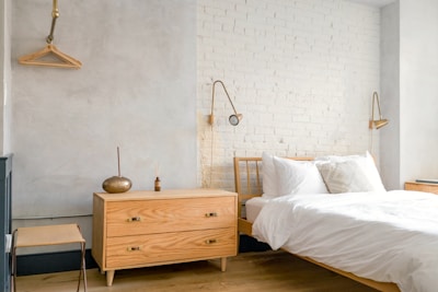A bright and minimalist bedroom with light-colored wooden furniture, including a bed and a chest of drawers. The walls are painted in soft gray and white brick, creating a calming atmosphere. Two modern wall-mounted lamps provide light, and a simple wooden coat hanger is visible on one wall.