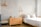 A bright and minimalist bedroom with light-colored wooden furniture, including a bed and a chest of drawers. The walls are painted in soft gray and white brick, creating a calming atmosphere. Two modern wall-mounted lamps provide light, and a simple wooden coat hanger is visible on one wall.