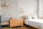 A bright and minimalist bedroom with light-colored wooden furniture, including a bed and a chest of drawers. The walls are painted in soft gray and white brick, creating a calming atmosphere. Two modern wall-mounted lamps provide light, and a simple wooden coat hanger is visible on one wall.