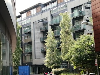 A modern, multi-story residential building with large glass balconies surrounded by lush green trees. The building has a sleek design with a mix of metal and glass surfaces. In the foreground, there is a blue dumpster and several trees providing some foliage.