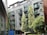 A modern, multi-story residential building with large glass balconies surrounded by lush green trees. The building has a sleek design with a mix of metal and glass surfaces. In the foreground, there is a blue dumpster and several trees providing some foliage.